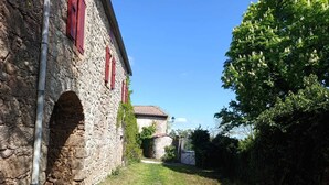 A stone building with red shutters, a narrow pathway, and a tree-lined background. - Authentic farmhouse at an elevation of 800 meters between Larzac and the Cévennes; sleeps 15 (Campestre-et-Luc)
