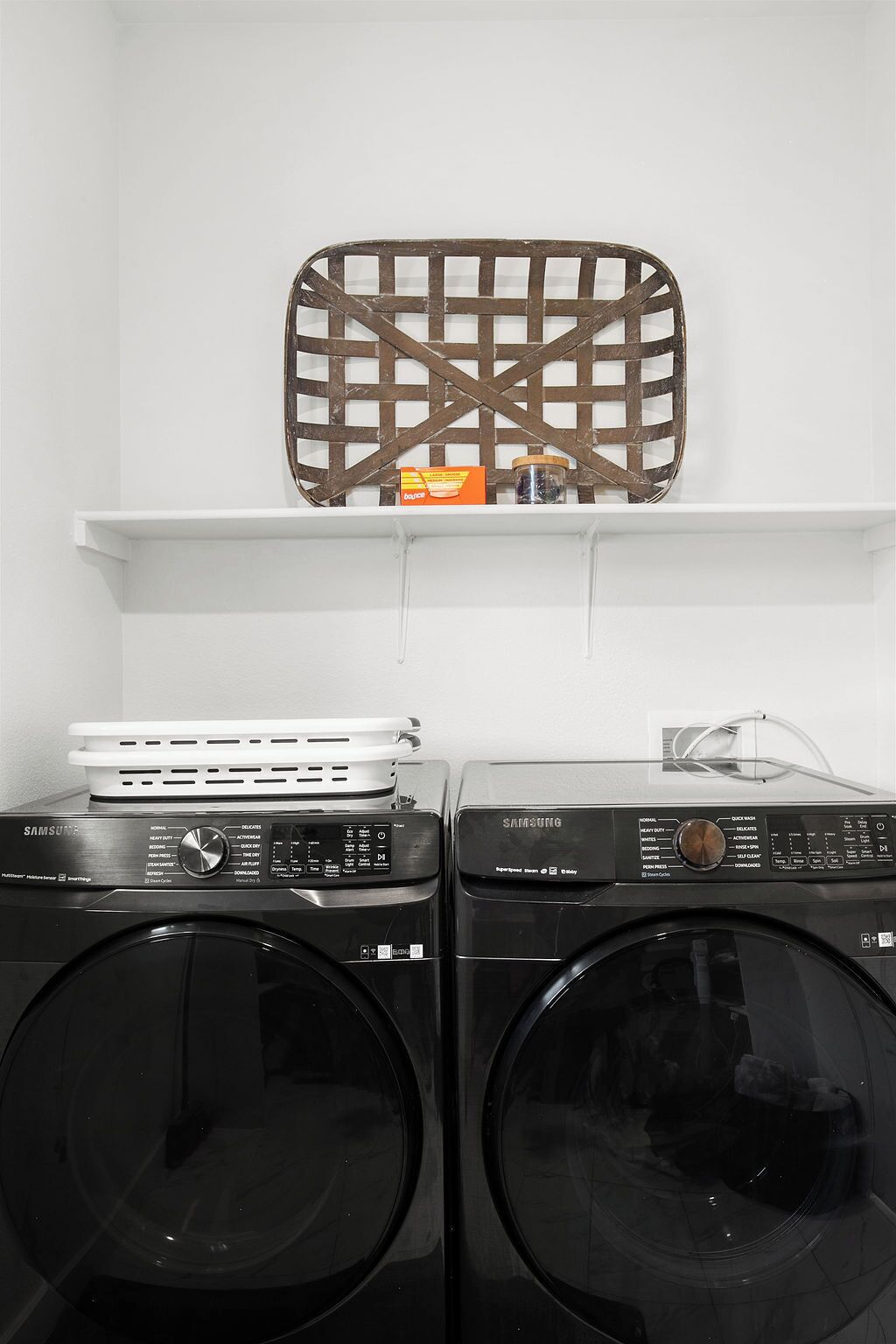 A laundry area with two front-loading washing machines, a shelf with a decorative metal piece, and a box of laundry detergent.