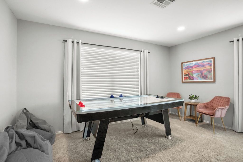 Quarto com mesa de futebol, uma pequena mesa com uma planta, duas cadeiras e uma janela com persianas.