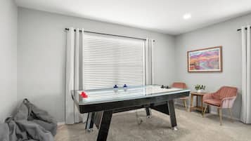 Quarto com mesa de futebol, uma pequena mesa com uma planta, duas cadeiras e uma janela com persianas.