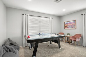 A room with a foosball table, a small table with a plant, two chairs, and a window with blinds.