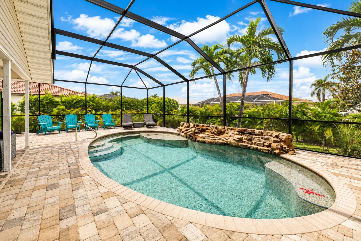 A swimming pool with a stone border, surrounded by a covered patio with chairs and a view of palm trees.