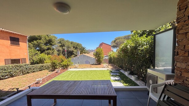 A patio with a table and chairs, a view of a garden, and a building in the background. - Apartment with garden in Saint-Raphaël (Saint-Raphaël)