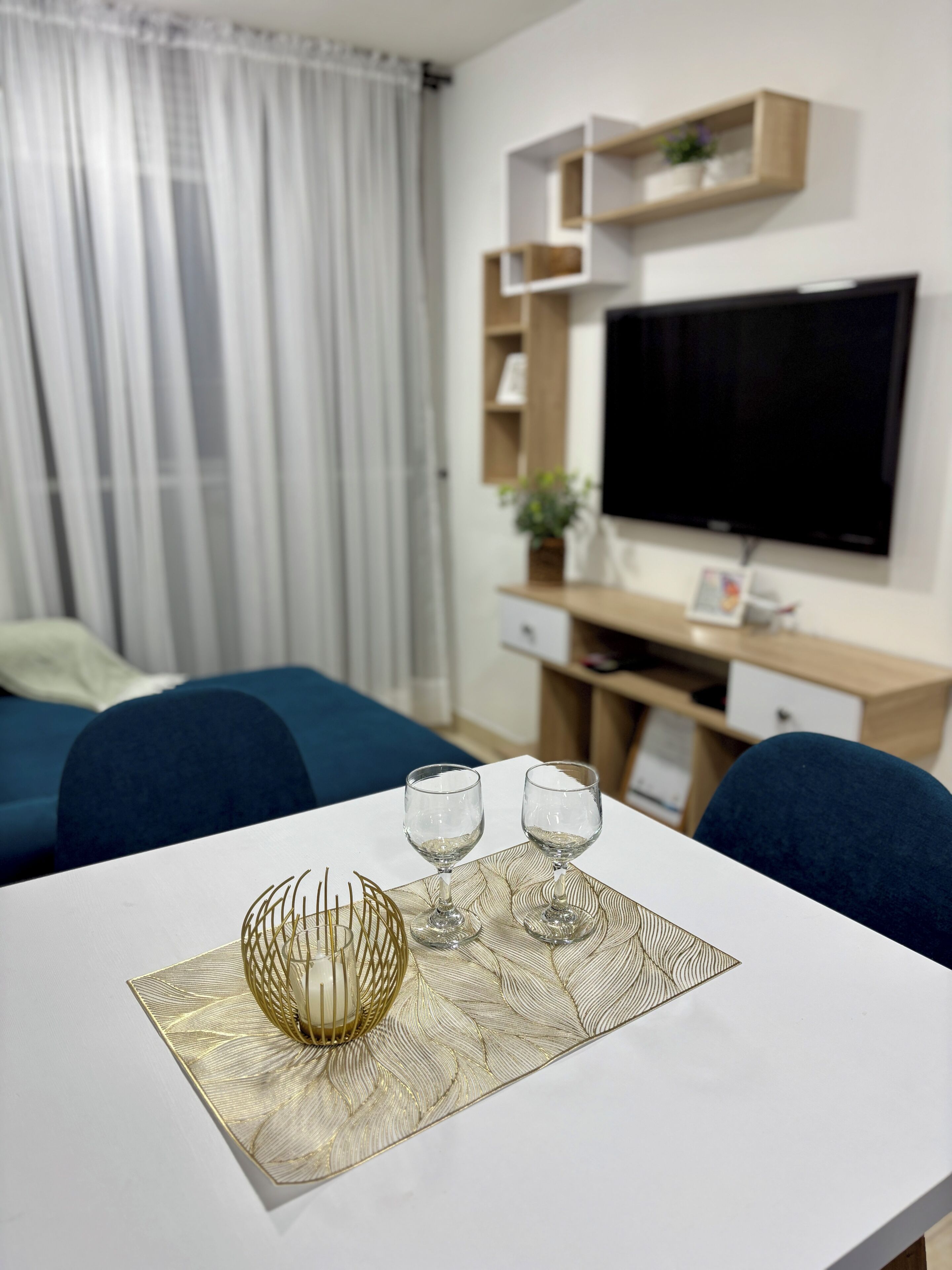 A modern living room with a white table, two glasses, a decorative centerpiece, a TV, and a blue sofa.