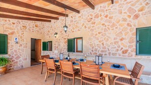 A stone-walled room with a wooden table and chairs, a hanging light fixture, and a door with green shutters. - Villa Ca Na Maria - By Dream Villas Pollensa (Pollensa)