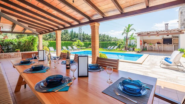 A wooden dining table set with plates, glasses, and cutlery, overlooking a swimming pool and outdoor seating area. - Villa Ca Na Maria - By Dream Villas Pollensa (Pollensa)