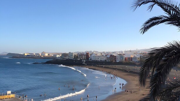 Una playa con un paseo marítimo, cielo despejado y un horizonte urbano distante.