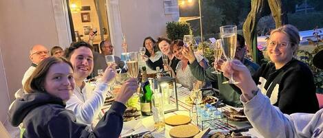 A group of people toasting at an outdoor table with food and drinks, with a building and trees in the background.