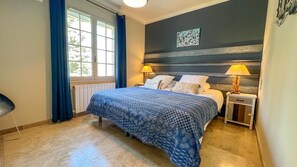 Une chambre à coucher avec un lit, deux tables de chevet, une fenêtre avec des rideaux et un mur orné d’un tableau.