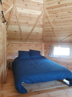 Een slaapkamer in een houten hut met een bed, kussens, een klein krukje en een raam.