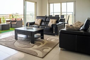 Un salon moderne avec un canapé en cuir noir, un fauteuil assorti, une table basse et une grande fenêtre offrant une vue sur l’extérieur.