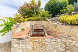 Un muro de piedra con zona de barbacoa, rodeado de plantas y árboles.