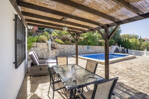 Un patio exterior con una mesa y sillas de vidrio, un muro de piedra y una piscina.