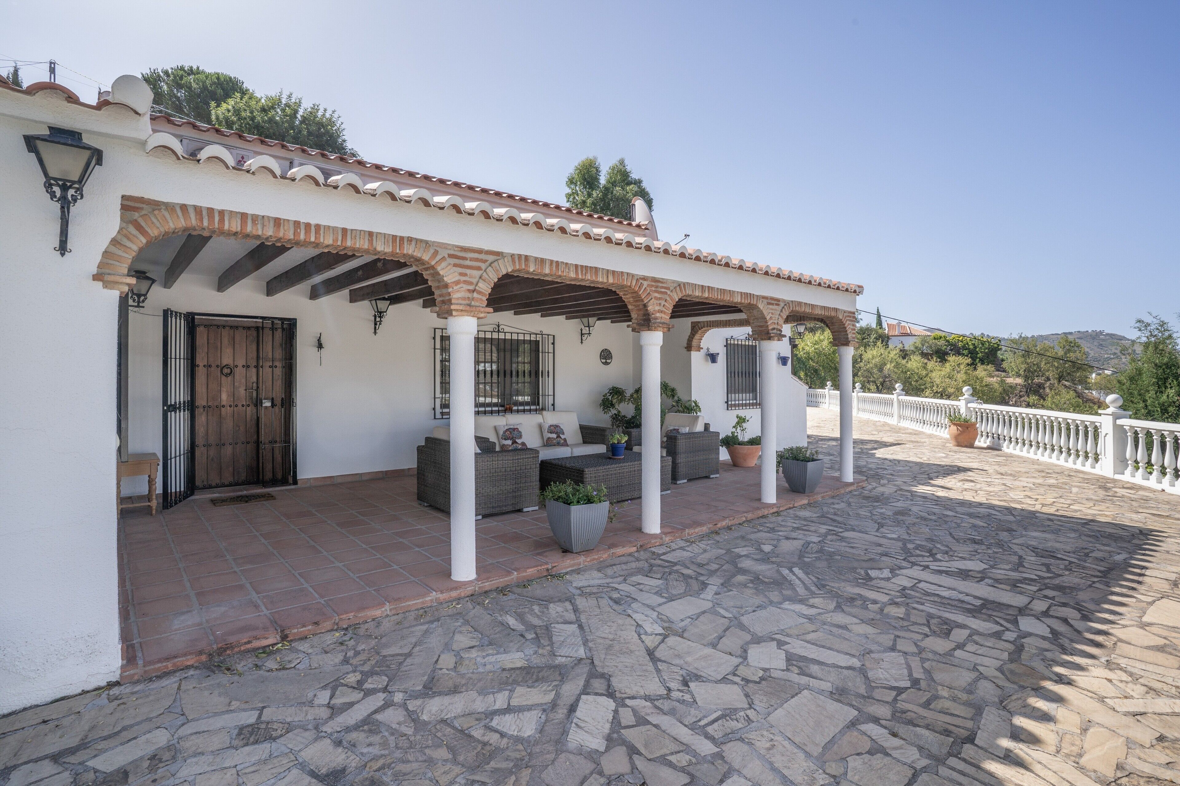 A covered outdoor patio with stone flooring, white pillars, and a white fence.