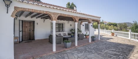 A covered outdoor patio with stone flooring, white pillars, and a white fence.