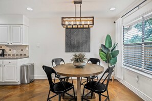 A dining area with a round wooden table, black chairs, a hanging light fixture, a potted plant, and a window with blinds. - Stylish Backyard Retreat BBQ Workspace (Austin)