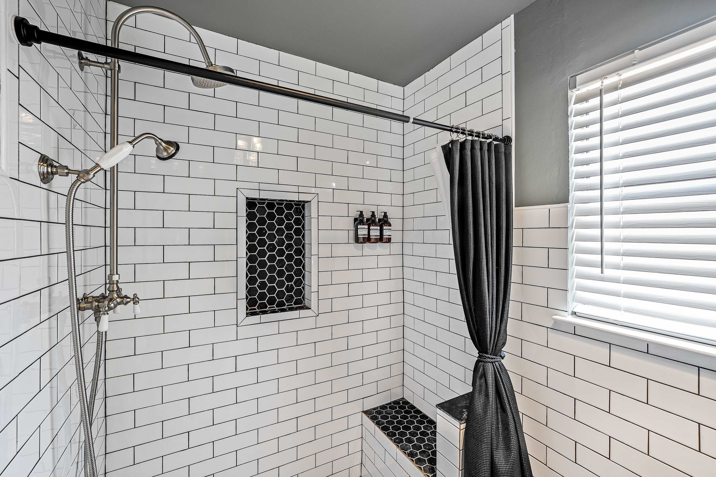 A shower with a curved showerhead, a handheld showerhead, and a window with blinds.