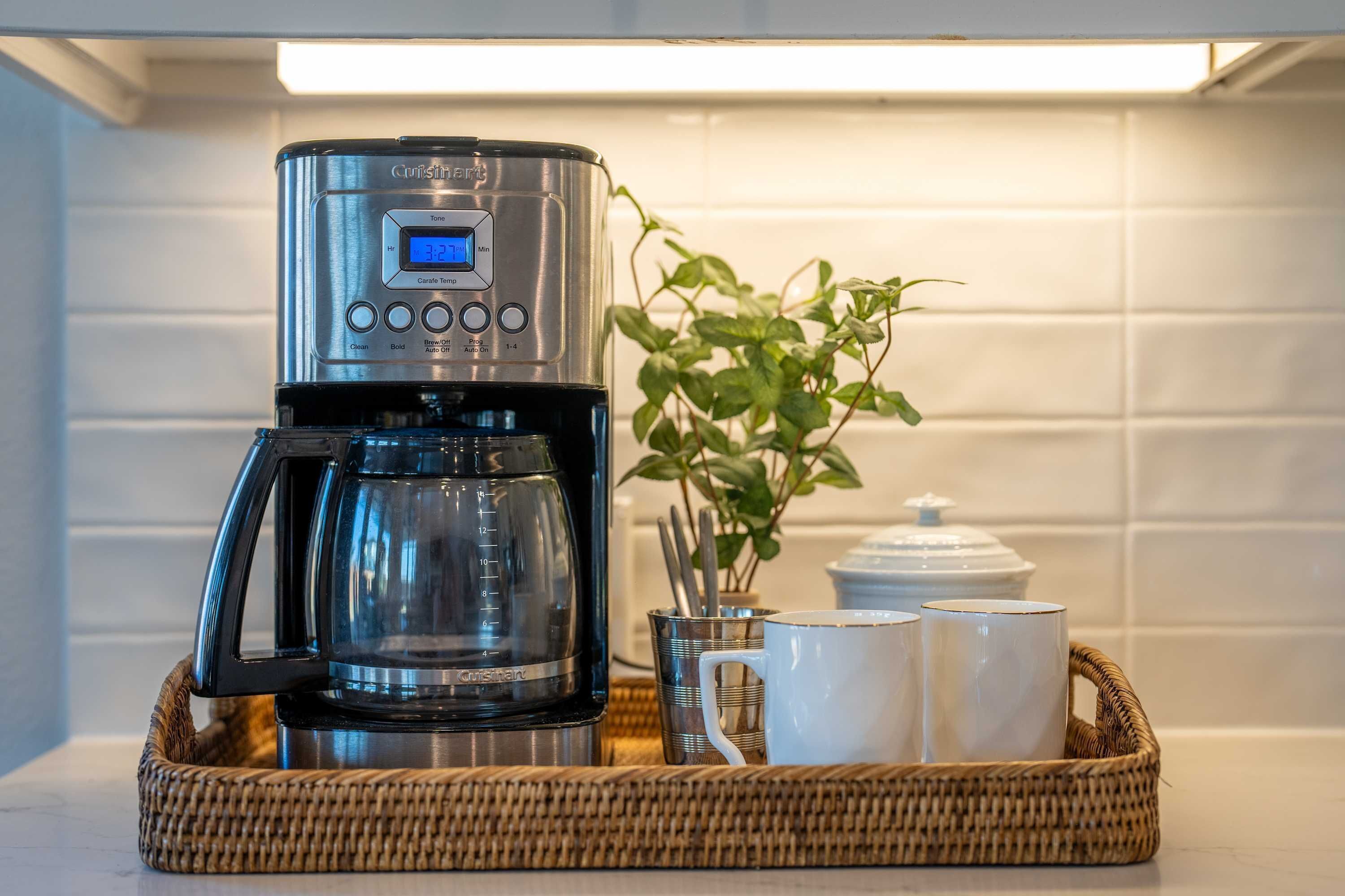 A coffee maker with a digital display, a glass carafe, two white mugs, and a wicker tray on a kitchen counter.
