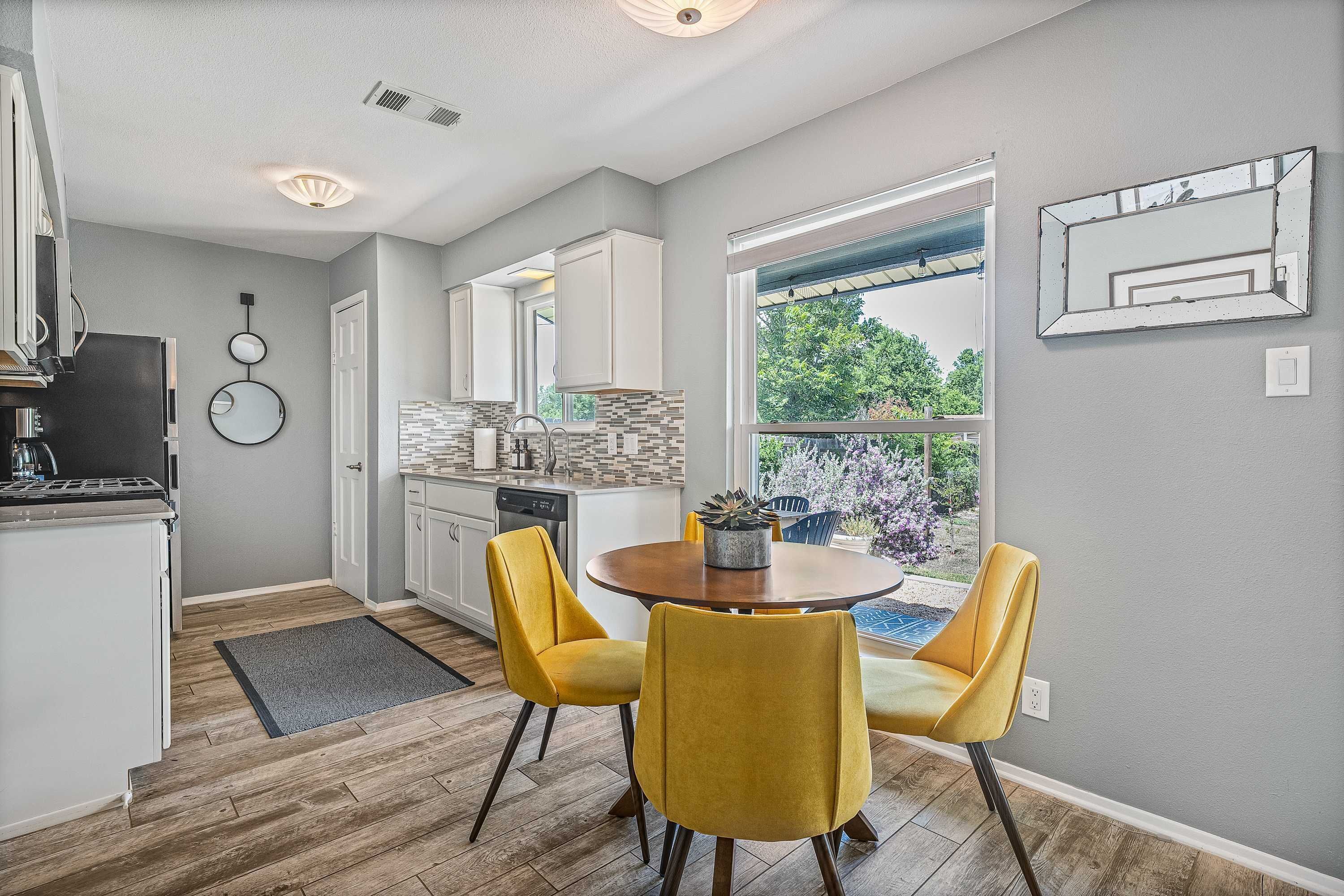 A modern kitchen with white cabinets, a dining area with yellow chairs, and a large window overlooking greenery.