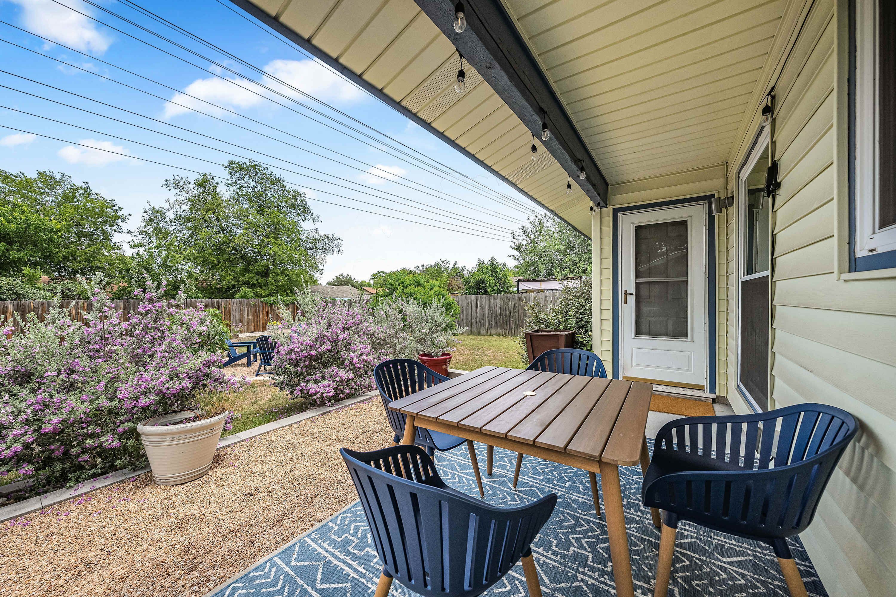 A covered patio with a wooden table and chairs, surrounded by potted plants and a garden.