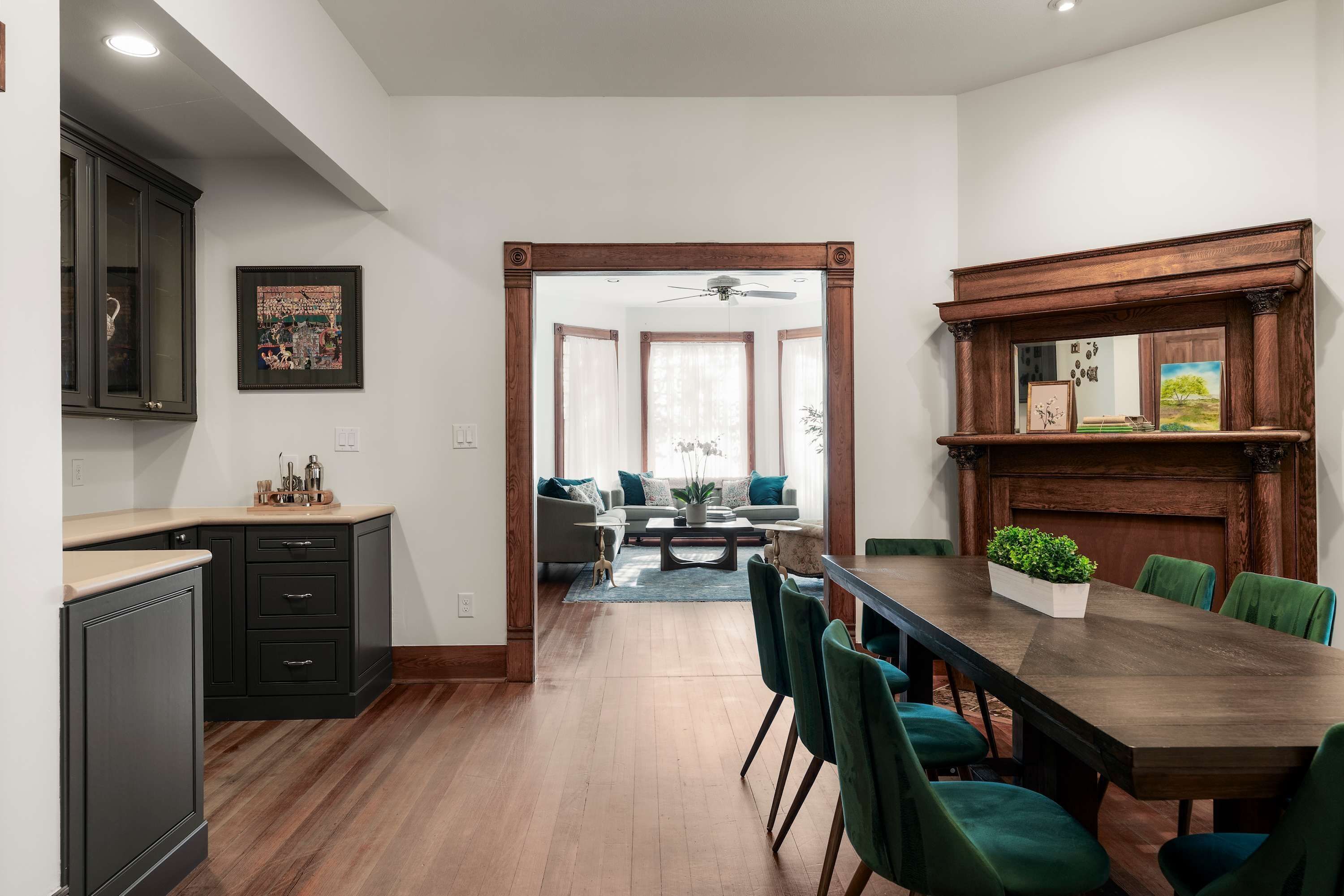 A modern kitchen with dark cabinetry, a dining area with a wooden table and green chairs, and a living room with a sofa and a large window.
