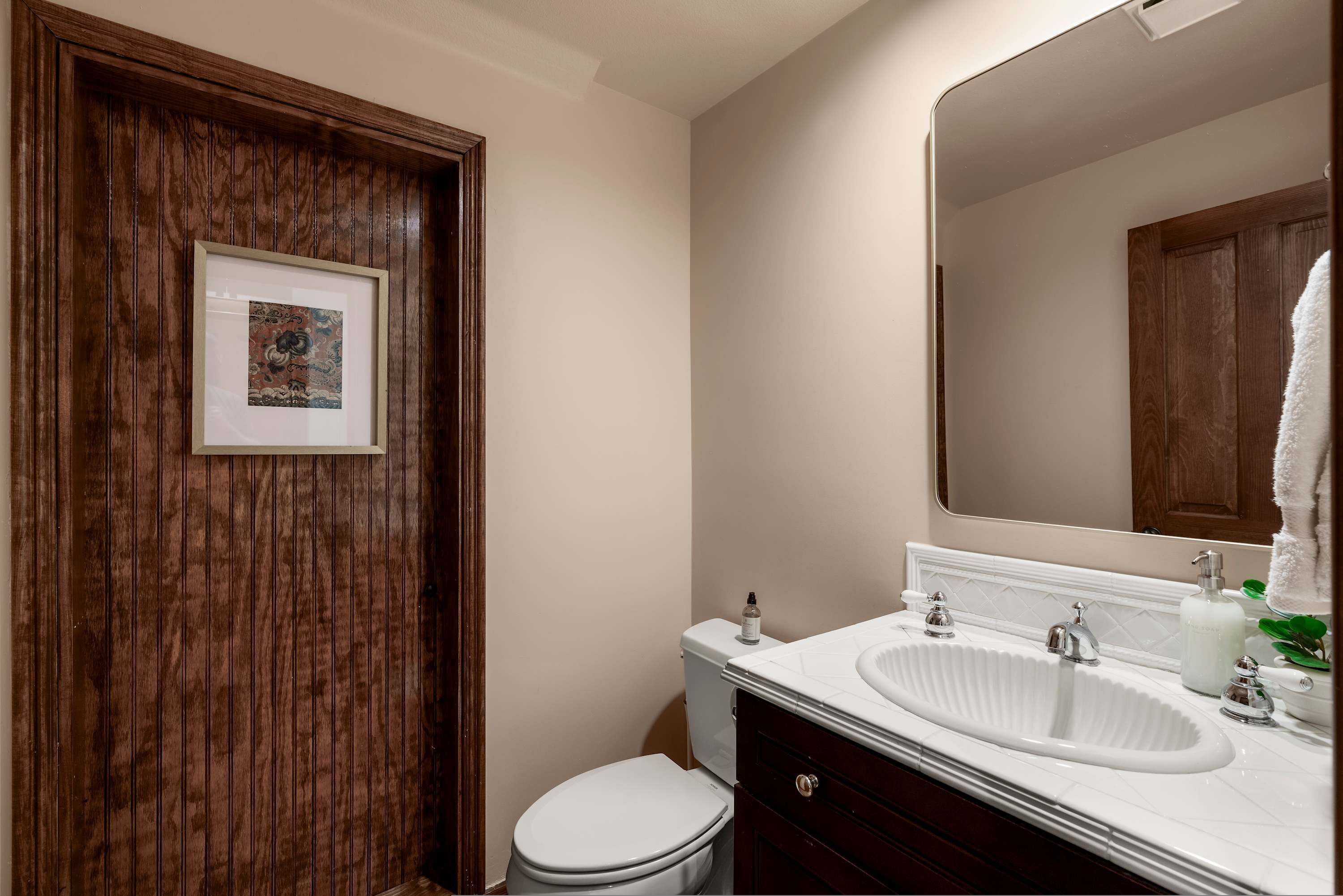 A bathroom with a wooden door, a white toilet, a sink with a mirror, and a framed picture on the wall.