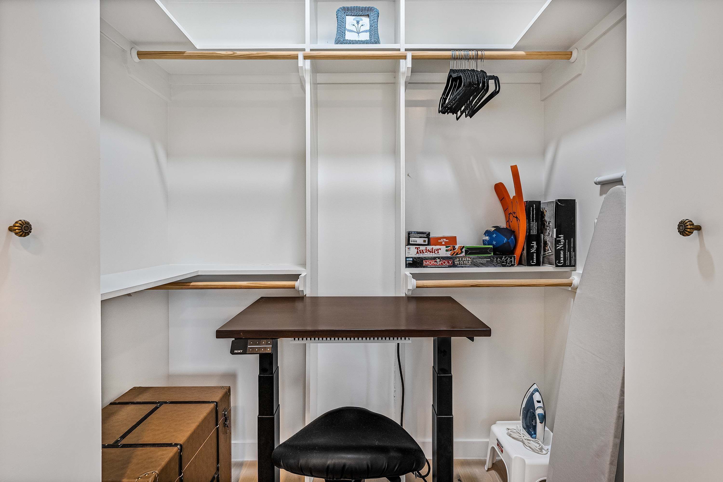 Quarto compacto com escrivaninha, cadeira, mesinha, prateleira com livros e uma caixa, e um cabideiro com roupas.