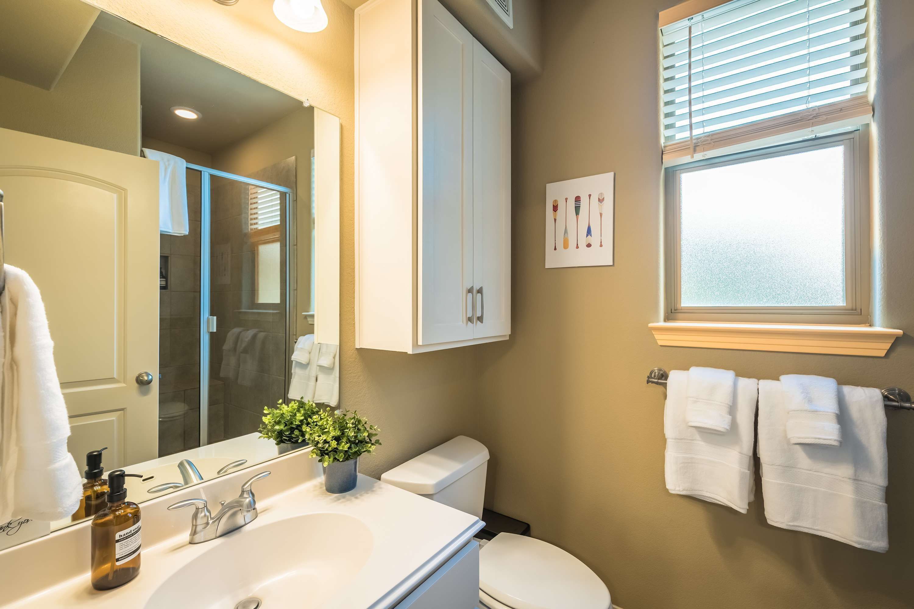 A bathroom with a white sink, a mirror, a cabinet, a toilet, and a window with blinds.