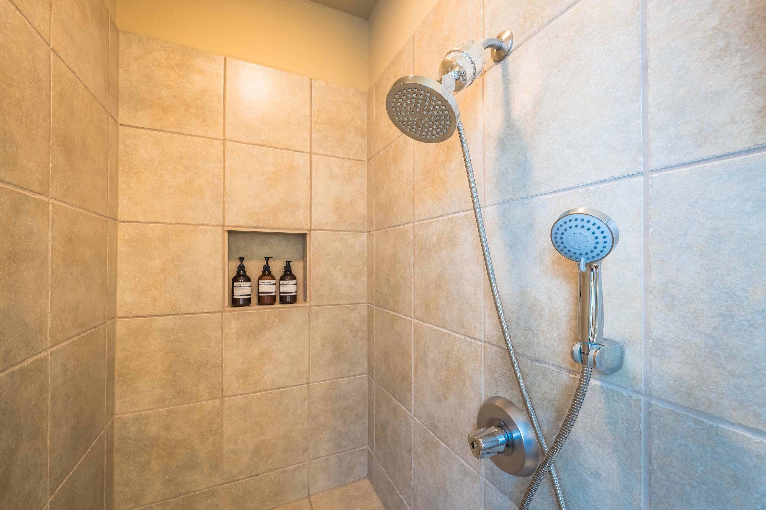 A shower with a handheld showerhead and a wall-mounted faucet, tiled walls, and a shelf with toiletries.