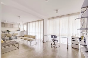 A modern kitchen with white cabinets, a dining area with wicker chairs, a glass desk, and a large window with sheer curtains.