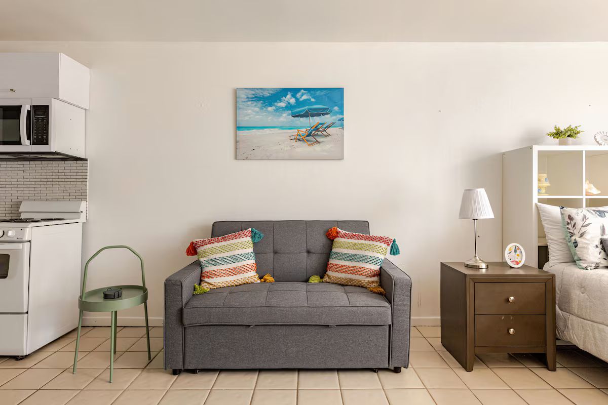 A living room with a grey sofa, colorful pillows, a painting of a beach scene, a small green chair, and a white kitchen area with a microwave.