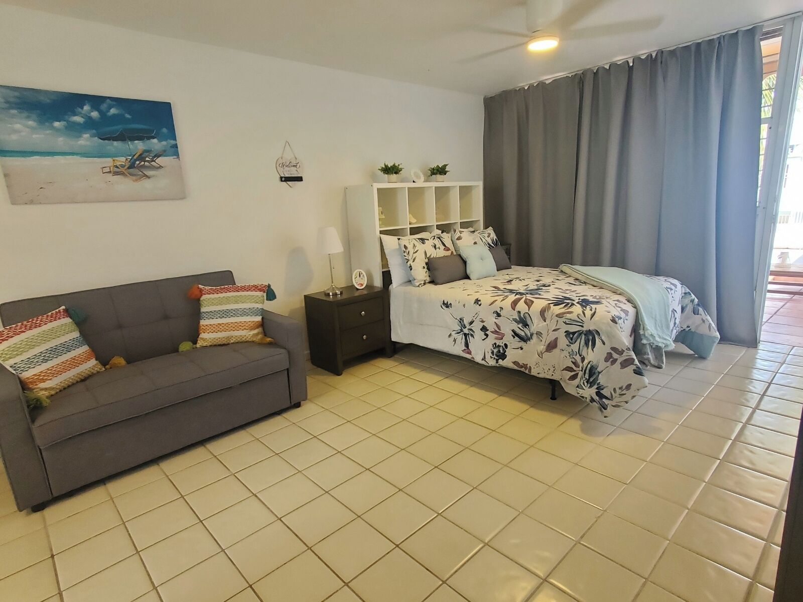 A bedroom with a bed, a sofa, a nightstand, a mirror, a shelf with plants, and a painting of a beach scene.