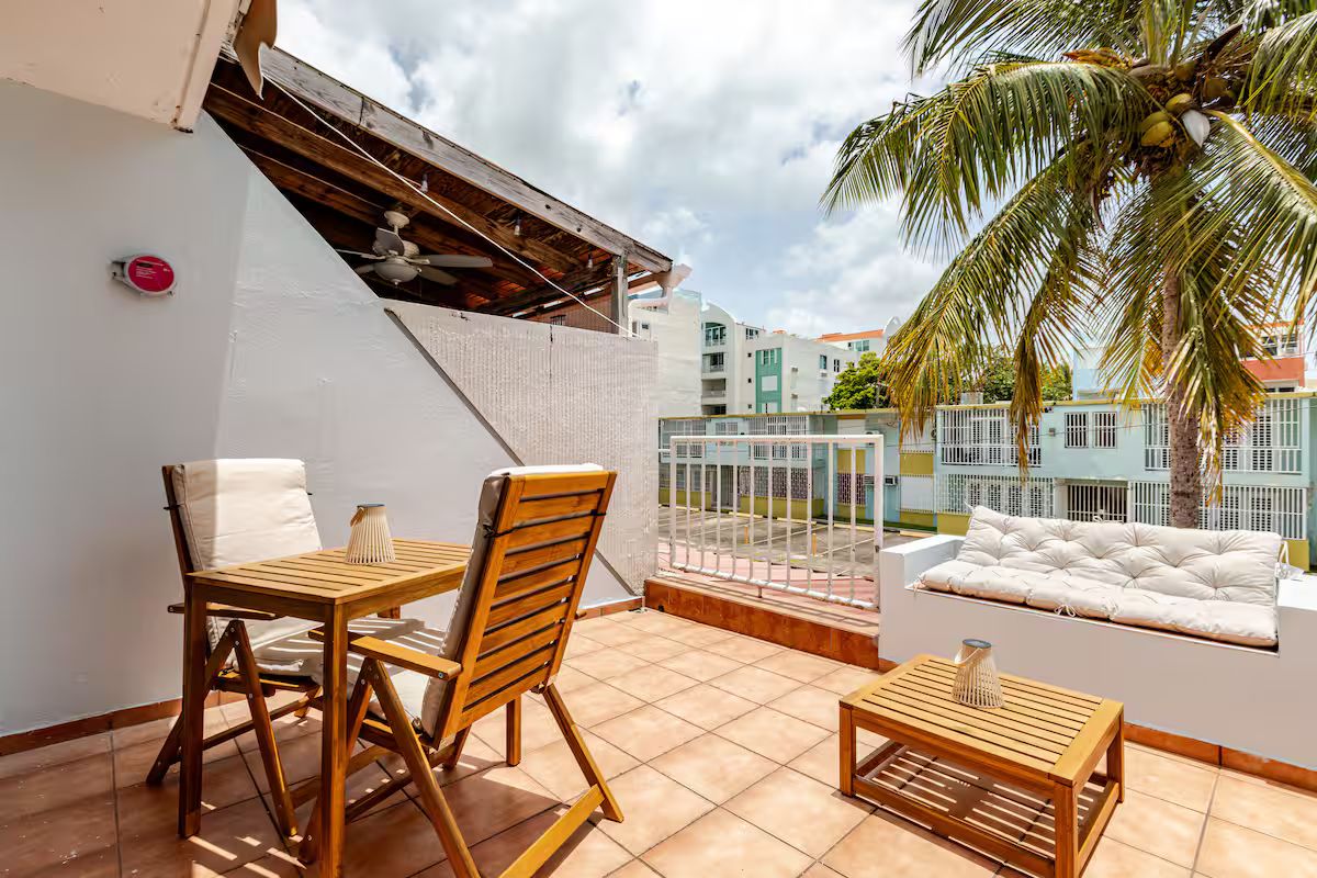 A balcony with a wooden table and chairs, a cushioned sofa, and a view of neighboring buildings.