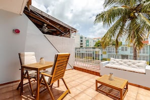 A balcony with a wooden table and chairs, a cushioned sofa, and a view of neighboring buildings. - Calm Cove Retreat a unique accommodation next to beautiful beaches Luquillo, PR. (Luquillo)