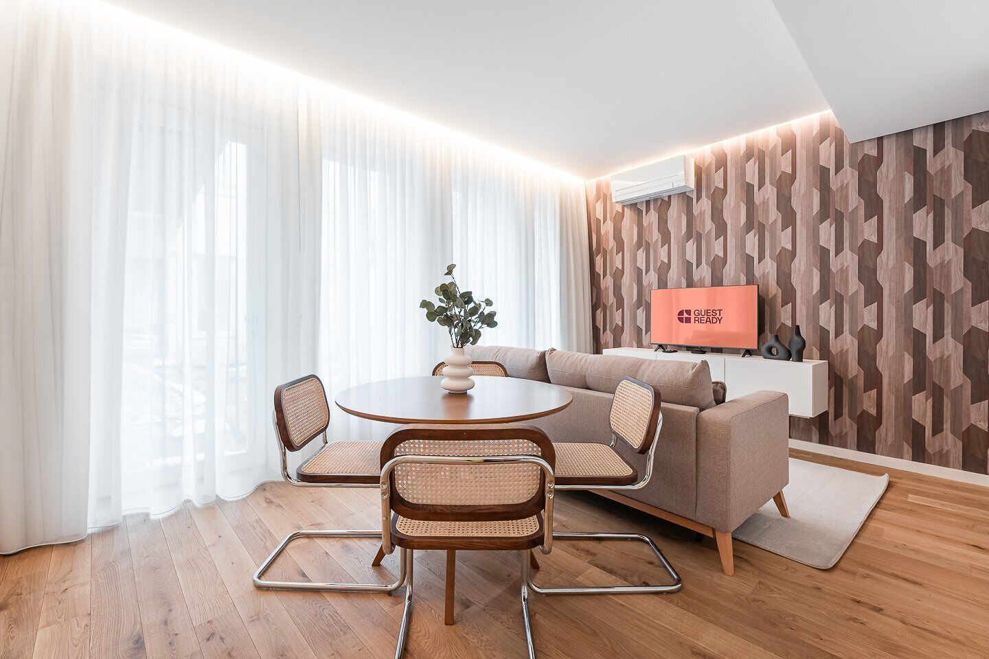 Ein modernes Hotelzimmer mit einem runden Esstisch, zwei Korbstühlen, einer Couch und einem Flachbildfernseher auf einer weißen Kommode.