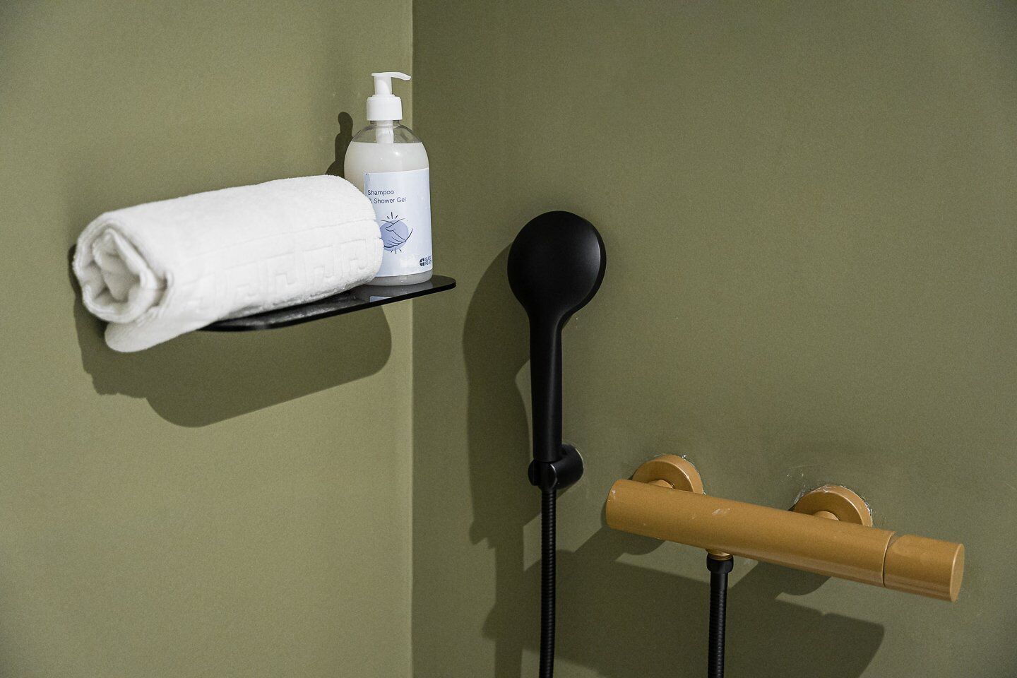 Une salle de bain avec une serviette posée sur une étagère, un flacon de shampoing, une pomme de douche noire et un porte-serviettes en bois.
