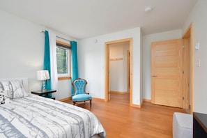 Quarto com cama, cadeira, mesa de cabeceira, janela com cortinas e porta de madeira.