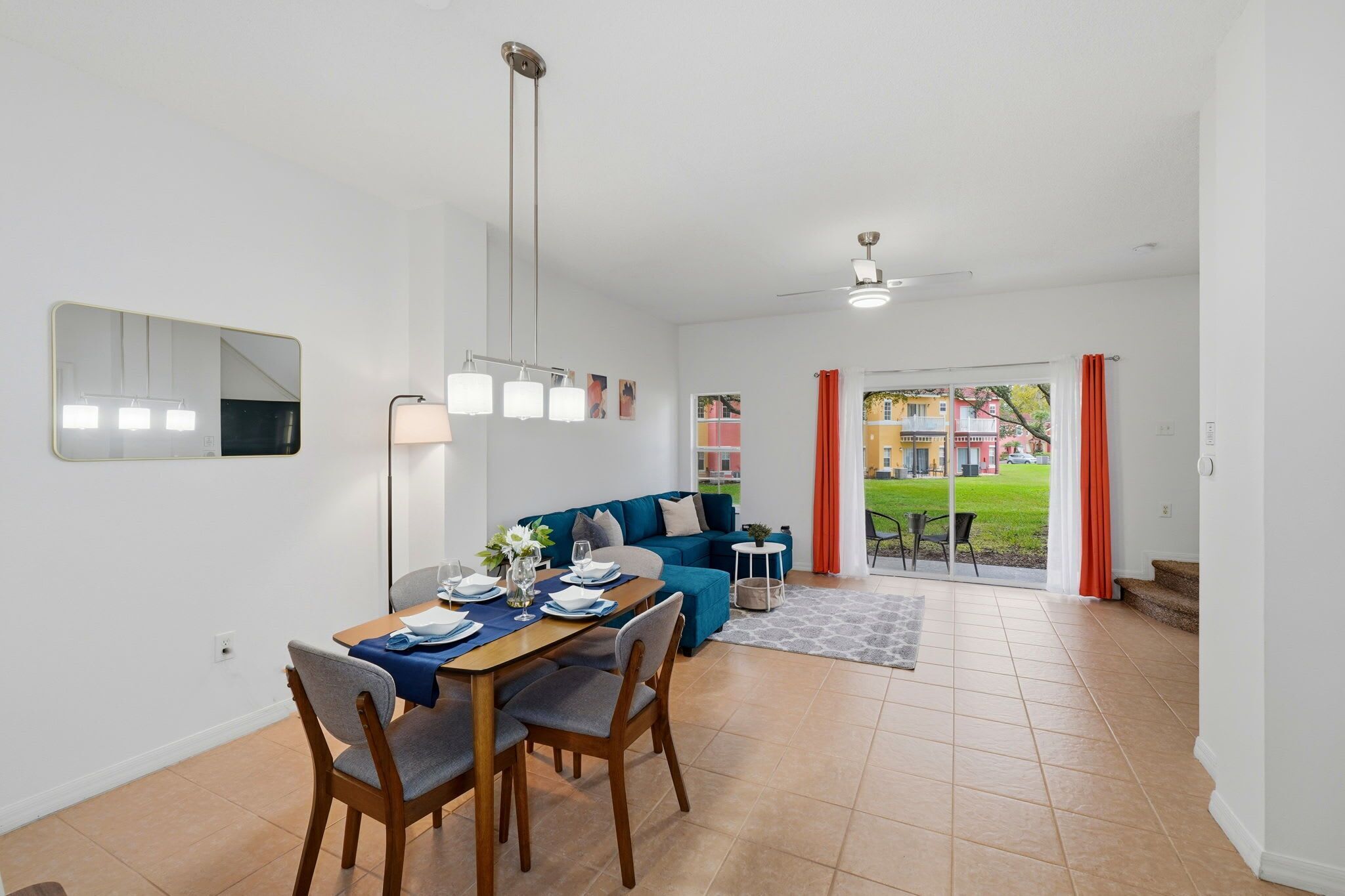 En spiseplads med et bord dækket til fire personer, en blå sofa og udsigt til et hus gennem en glasdør.