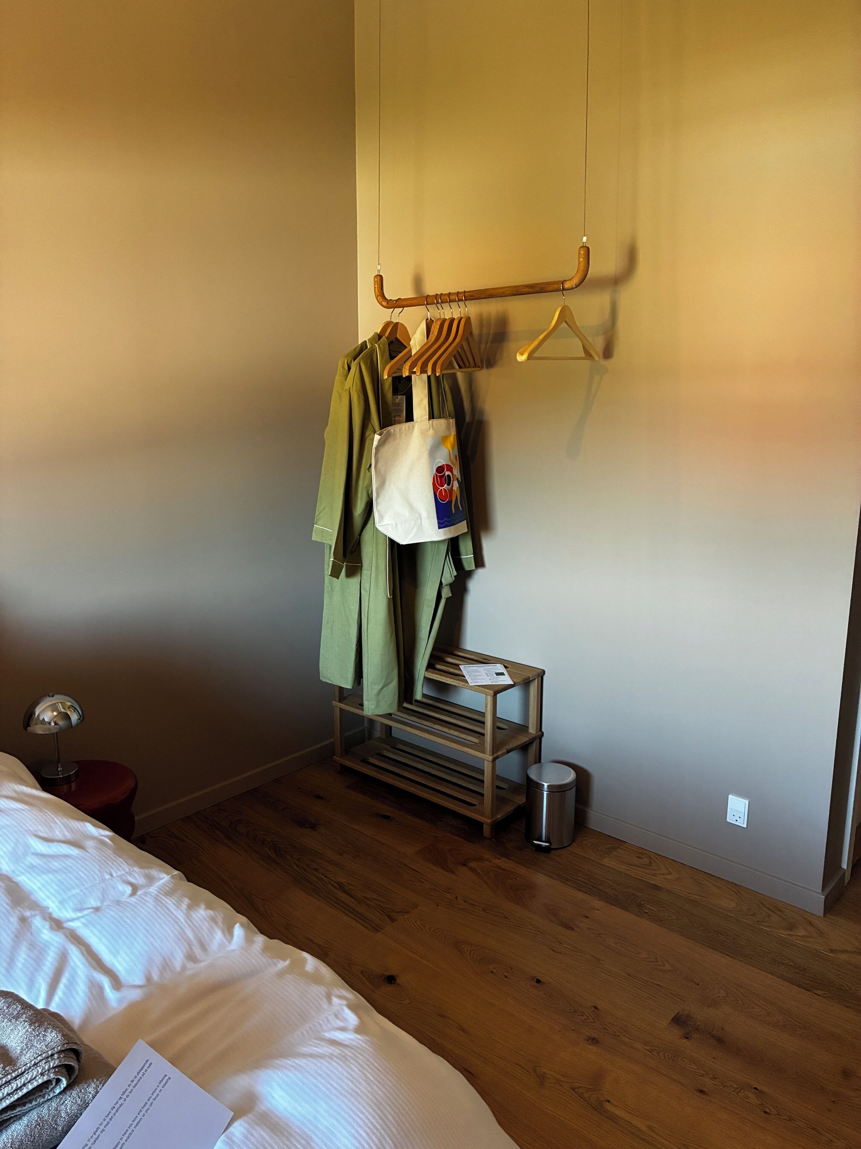 Hotelkamer met een bed, een houten vloer, een kapstok, een tafeltje met een tas en een prullenbak.