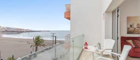 Een balkon met glazen hekwerk met uitzicht op een strand, een tafeltje met stoelen en een rode lantaarn.