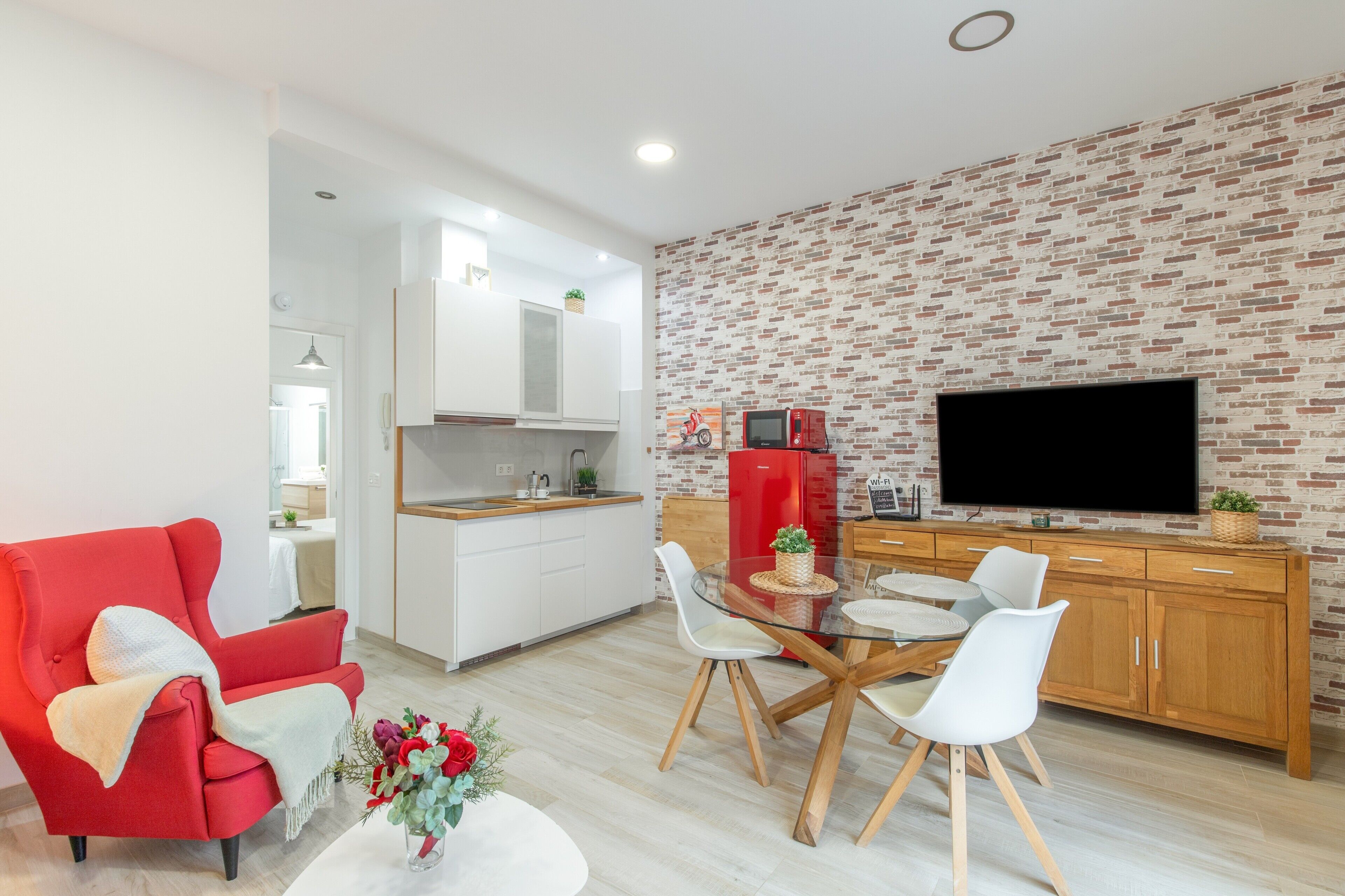 Una sala de estar moderna con un sillón rojo, una mesa de comedor de vidrio y una zona de cocina con pared de ladrillo y televisor de pantalla plana.