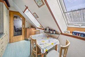 A dining area with a table set for two, a wooden chair, a small shelf with kitchen items, and a window with a view.