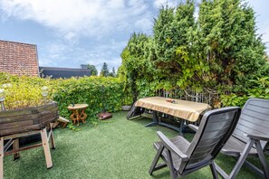 Un patio al aire libre con una mesa y sillas, rodeado de abundante follaje y un muro de ladrillo.