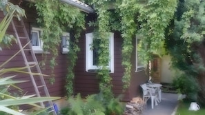 Una casa con fachada de madera, una ventana y una puerta blanca. Hay una silla blanca y una mesa afuera.