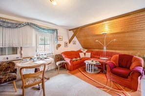 Una sala de estar acogedora con techo de madera, un sofá rojo, un sillón estampado, una mesa de comedor con mantel de encaje y una lámpara de pie.