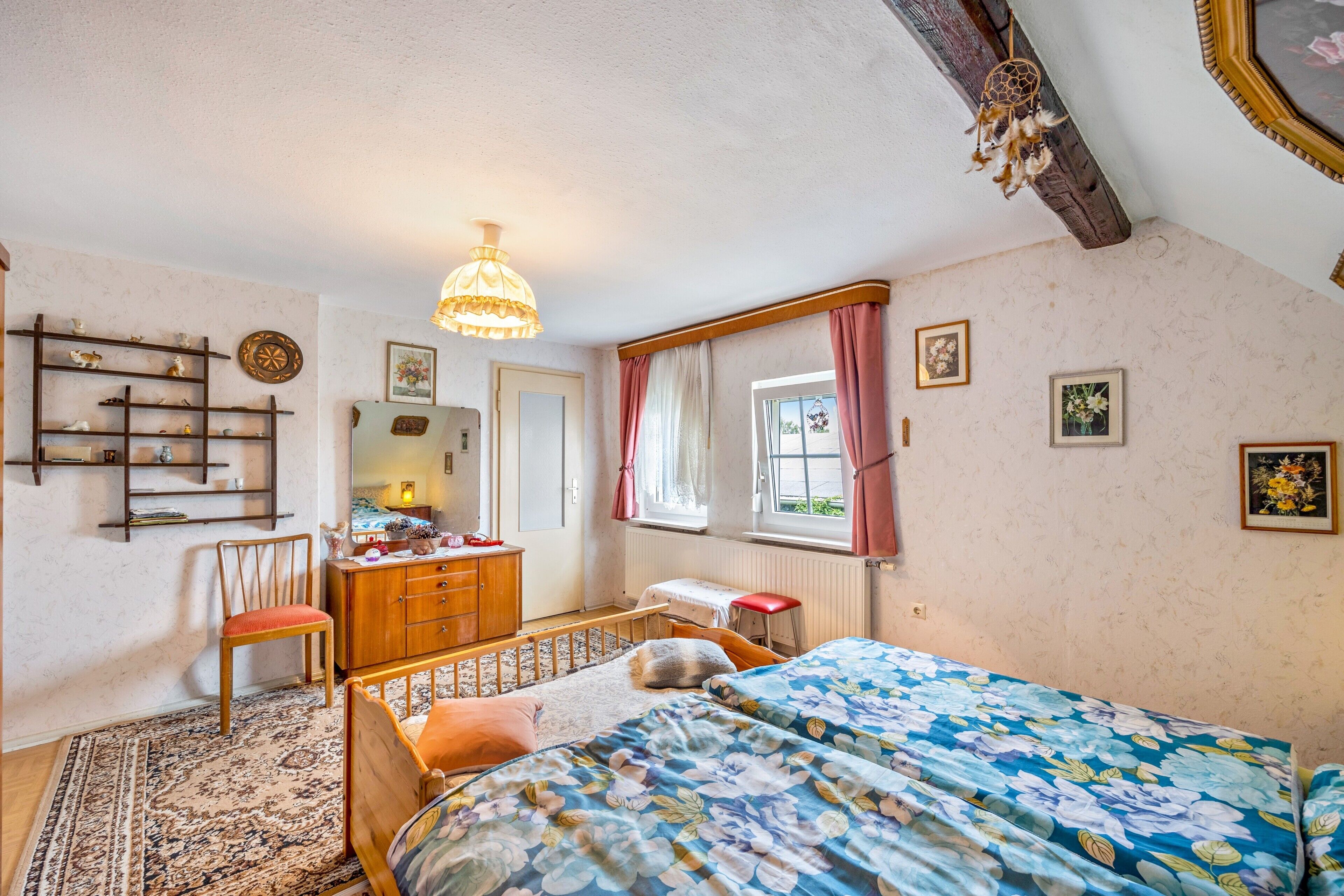 Une chambre à coucher comprenant un lit, une commode, une chaise, un bureau, une fenêtre avec des rideaux et une étagère fixée au mur, ornée d’objets décoratifs.