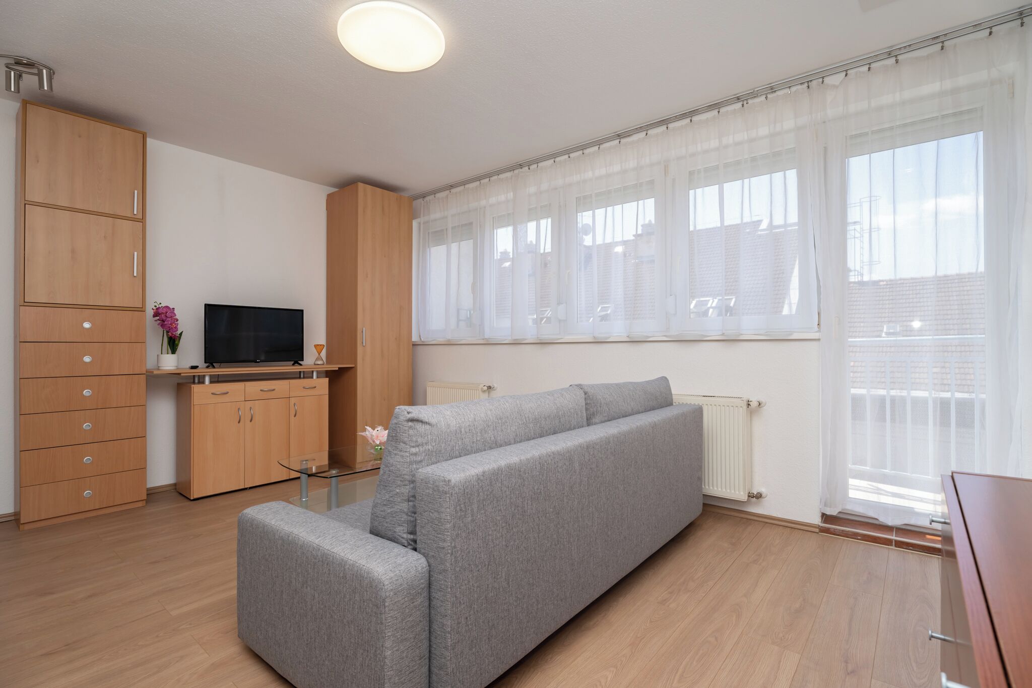 Una sala de estar moderna con un sofá gris, armarios de madera, un televisor de pantalla plana y ventanas grandes con cortinas translúcidas.