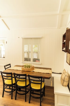 A dining area with a wooden table, four chairs, a basket with flowers, and a window with blinds. - Charming, Vintage & Authentic Getaway - Sunroom - Free Parking! (Forest Falls)