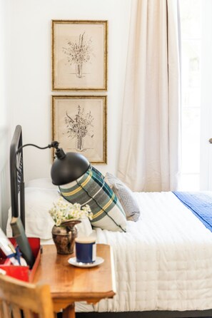 A bedroom with a bed, a desk with a lamp, a vase with flowers, and two framed pictures on the wall. - Charming, Vintage & Authentic Getaway - Sunroom - Free Parking! (Forest Falls)