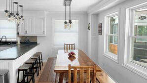 Een keuken met witte kastjes, een houten eettafel en een bankje. Er hangen lampen en er is een raam met jaloezieën.